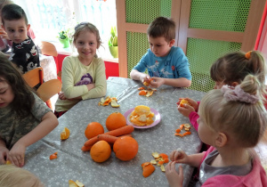 Piecioro dzieci siedzi przy stoliku i obiera pomarańcze. Na środku stołu leżą marchewki i pomarańcze – zarówno obrane jak i nieobrane.
